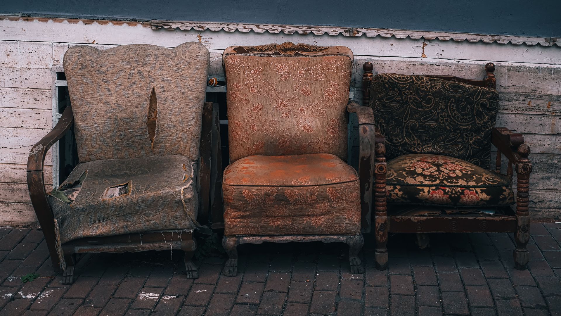 Three old, worn-out vintage chairs on a brick pathway against a wooden wall.
